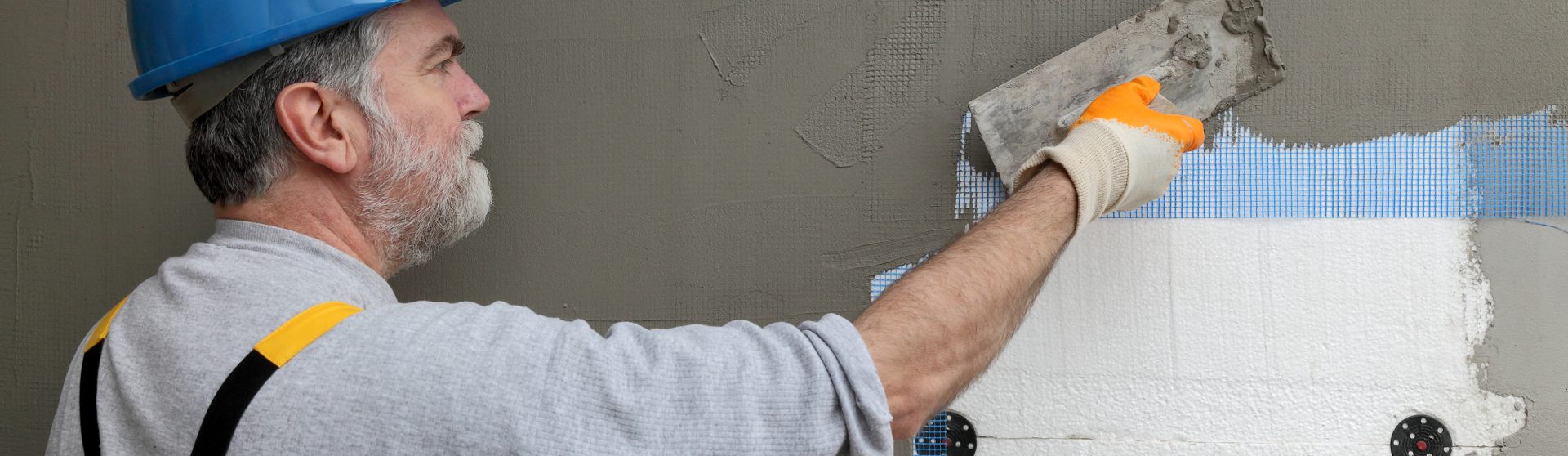 Männliche Person mit blauem Schutzhelm verputzt mit einer Kelle eine gedämmte Aussenwand