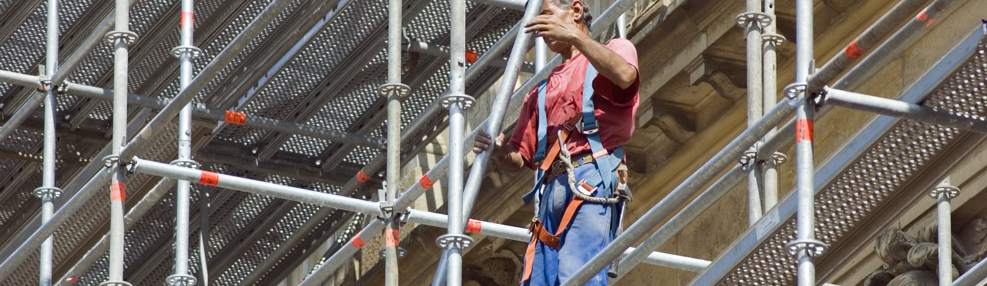Person in rotem T-Shirt und Latzhose steht auf einem Gerüst und montiert Gerüstteile