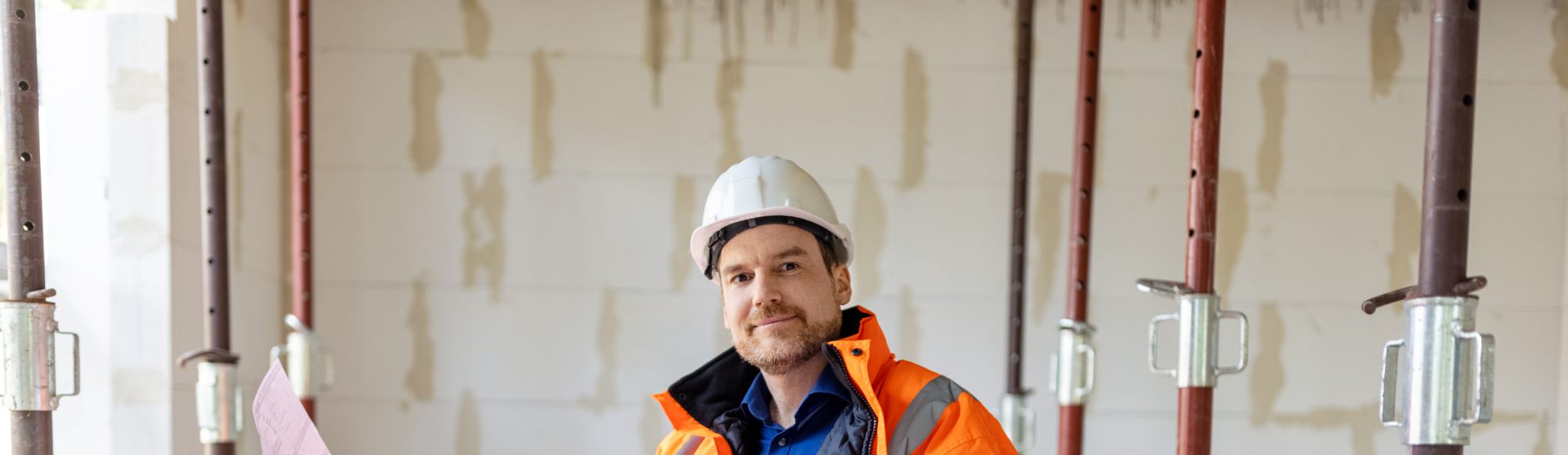 Man in Sicherheitskleidung steht in einer Baustelle mit Deckensicherungselementen. Er hält einen Plan in der Hand und schaut in die Kamera.
